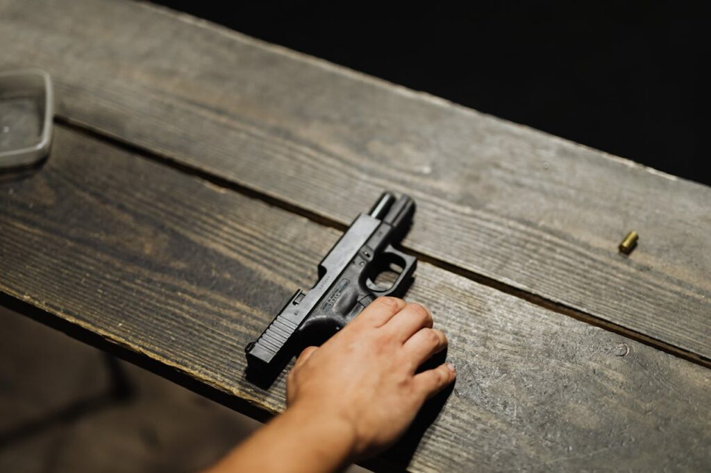 pexels-photo-5202395 Close-up of a hand holding a handgun on a wooden table with bullet casing.